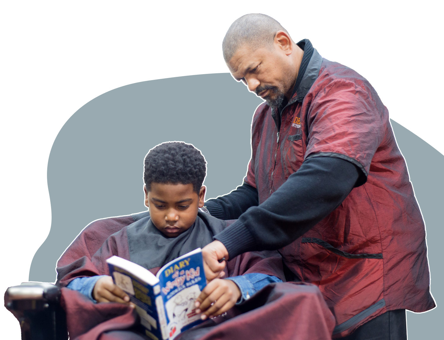 Black boy reading a book while getting his hair cut by a Black barber.