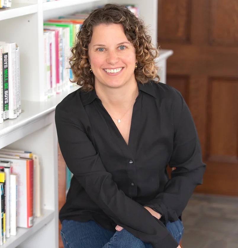 Picture of Dr. Molly Ness in a black shirt. She's sitting in front of a white bookshelf filled with books.