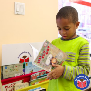 Black boy opening a book called Fly Guy! in front of a Barbershop Books bookshelf.